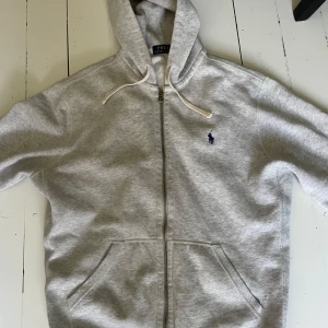 Grå hoodie från Polo Ralph Lauren - Snygg grå hoodie från Polo Ralph Lauren med dragkedja och klassisk broderad logga på bröstet. Tröjan har huva med snörning och två fickor framtill. Perfekt för en avslappnad stil. Storlek L. Pris kan diskuteras vid snabb affär…
