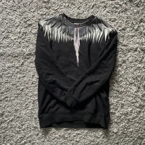 Marcelo burlon sweatshirt - Säljer en svart sweatshirt från Marcelo Burlon County of Milan med ikoniskt vingmönster i grått och vitt över axlar och bröst. Tröjan är i mjuk bomull och har rund hals samt långa ärmar. Perfekt för dig som vill sticka ut med en unik design.