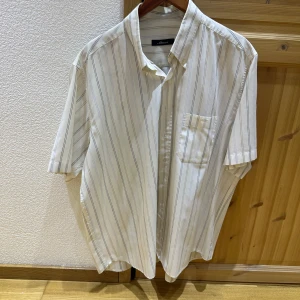 Beige och blå randig skjorta från UNIQLO - Säljer en klassisk beige skjorta med tunna blå ränder. Skjortan har korta ärmar, bröstficka och en avslappnad passform. Perfekt för en stilren och enkel look.