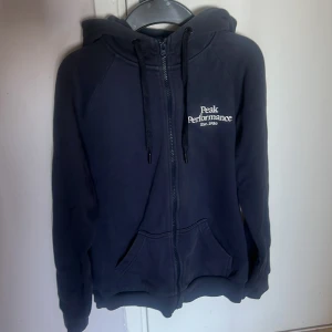 Mörkblå hoodie från Peak Performance - Mysig mörkblå hoodie från Peak Performance med dragkedja och huva. Har två fickor framtill och ribbade muddar vid ärmslut och nederkant. Perfekt för kyliga dagar eller som ett extra lager.