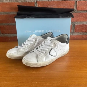 Vita sneakers från Philippe Model - Säljer ett par vita sneakers från Philippe Model med svarta detaljer vid hälen och på sidan. Skorna har klassisk snörning och en platt sula. Perfekta för en stilren och avslappnad look.