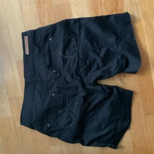 Svarta shorts y2k - Svarta shorts med dekorativa nitar på bakfickorna och tydliga sömmar. Knappar framtill med stjärndetaljer. Klassisk femficksmodell i bomull. Likt y2k och mycket snygga.