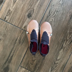Nike Mercurial PRO fotbollsskor i koppar och svart - Säljer ett par Nike Mercurial PRO fotbollsskor i kopparfärg med svarta detaljer. Skorna har en strumpliknande passform utan traditionell snörning så att man inte behöver knyta skorna. 