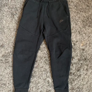 Svarta mjukisbyxor från Nike - Svarta mjukisbyxor från Nike med dragsko i midjan och diskret logga på vänster ben. Byxorna har normal passform, muddar vid bensluten och praktiska fickor, varav en med dragkedja på baksidan.