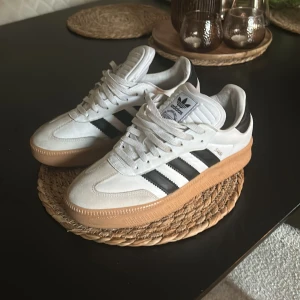 Adidas Samba OG sneakers med gummisula - Säljer ett par Adidas Samba OG sneakers i vitt skinn med svarta ränder och detaljer. Skorna har klassisk gummisula och rund tå. Perfekta för dig som gillar retrostil och streetwear. Ikonisk Adidas-logga på plösen och svart hälparti.