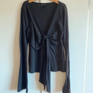 Svart omlottopp med knyt från Gina Tricot - Svart långärmad omlottopp från Gina Tricot med djup v-ringning och knyt framtill. Perfekt att styla med jeans eller kjol för en trendig look.