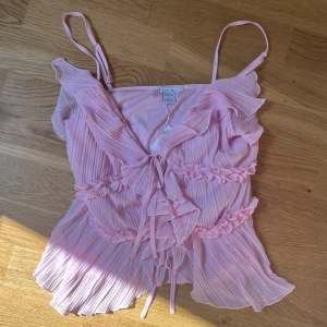 Ljusrosa volanglinne volangblus med knyt - Supersöt ljusrosa blus  med volanger och knyt framtill. Plisserad design och tunna axelband ger en romantisk känsla. Perfekt för vår och sommar!Söt trendig ljusrosa volang linne  NY MED PRISLAPP!!  Är storlek L men väldigt liten i storleken så passar S