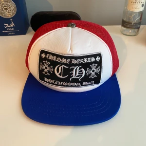 Chrome Hearts keps i rött, vitt och blått - Säljer en Chrome Hearts truckerkeps med vit front, röd mesh och blå skärm. Kepsen har en broderad svartvit patch framtill med texten 'Chrome Hearts Hollywood, USA' och justerbar snapback baktill.