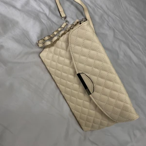 Beige quiltad axelväska - Snygg beige axelväska med quiltat mönster och kedjedetaljer på axelremmen. Väskan har en elegant design med en praktisk stängning framtill. Perfekt för att ge en stilren touch till din outfit.