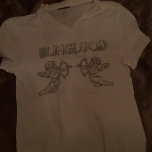 Vit t-shirt med bling och änglar - Säljer en vit t-shirt med glittrande text 'BLINGLUND' och två änglar i strass på framsidan. T-shirten har en v-ringad hals och korta ärmar. Perfekt för dig som gillar att sticka ut med lite extra bling! ✨