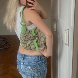 Beige och grön axelväska med bokstäver - Trendig axelväska i beige med gröna detaljer och stora bokstäver. Väskan har en unik design med en cirkulär metallring framtill och gröna axelremmar. Perfekt för att ge din outfit en färgklick!