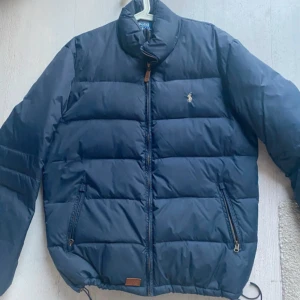 Mörkblå dunjacka från Polo Ralph Lauren - Säljer en snygg mörkblå dunjacka från Polo Ralph Lauren med dragkedja och broderad logga på bröstet. Jackan har långa ärmar och praktiska fickor med dragkedja. Perfekt för kyliga dagar! 💙
