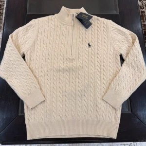 Beige stickad tröja från Polo Ralph Lauren - Säljer en stilren beige stickad tröja från Polo Ralph Lauren med kabelstickat mönster och halv dragkedja. Tröjan har långa ärmar och en liten broderad logga på bröstet. Perfekt för kyliga dagar!