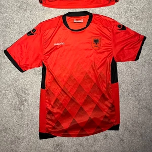 Albanien fotbollströja hemma 2017 - Säljer en röd fotbollströja från Macron med svarta detaljer och ett emblem på bröstet. Tröjan har korta ärmar och är tillverkad i 100% mikrofiber. Perfekt för fotbollsfans som vill visa sitt stöd.