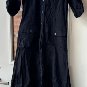 Svart långklänning med knappar - Snygg svart långklänning med knappar framtill och praktiska fickor. Perfekt för en stilren look. Klänningen har en klassisk design som passar till många tillfällen.