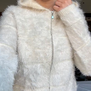 Vit fluffig kofta med dragkedja och luva  - Mysig vit kofta med fluffig textur och dragkedja framtill. Perfekt för kyliga dagar med sin långa ärmar och bekväma passform. Jackan har en huva som ger extra värme och stil.