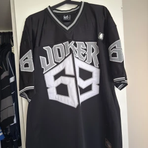 Joker brand jersey xxxl - Säljer en svart jersey från Joker med grå detaljer och texten 'Joker 69' på både fram- och baksidan. Tröjan har korta ärmar och en v-ringad hals med randiga muddar. Perfekt för en avslappnad stil.