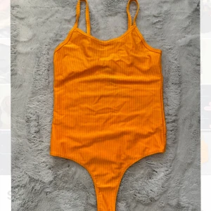 Orange ribbad bodysuit - Säljer en snygg orange ribbad bodysuit med smala axelband. Perfekt för att styla med jeans eller kjol. Den har en enkel och stilren design som passar till många tillfällen.