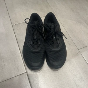 Svarta sneakers från Nike - Snygga svarta sneakers från Nike med snörning. Skorna har en stilren design med en ovandel i syntetmaterial och en bekväm sula. Perfekta för både träning och vardag. Finns lite vita färgfläckar