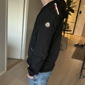 Svart jacka från Moncler  - Säljer en snygg svart jacka från Moncler med huva och dragkedja. Jackan har Moncler-logga på ärmen och ränder i rött, vitt och blått på huvan. Perfekt för en stilren look. (Från pandabuy)