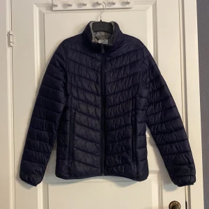 Mörkblå dunjacka från McKinley - Säljer en stilren mörkblå vårjacka från McKinley. Jackan har en quiltad design med dragkedja framtill och hög krage för extra värme. Perfekt för kyliga dagar och enkel att matcha med olika outfits. Ingen huva.