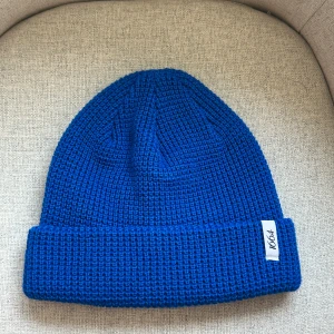 Blå stickad mössa  - Snygg blå stickad mössa, perfekt för kyliga dagar. Tillverkad av 50% återvunnen polyester och 50% akryl. Mössan har en bred uppvikt kant och en diskret etikett på sidan.