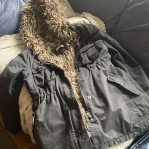 Svart parkajacka med fuskpäls - Säljer en svart parkajacka med fuskpäls i luvan. Jackan har dragkedja och knappar framtill samt flera praktiska fickor. Perfekt för kyliga dagar.