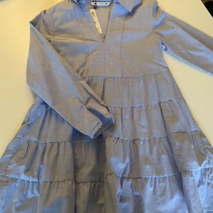 Blå klänning från Zara - Säljer en söt blå klänning från Zara med volanger och v-ringning. Klänningen har långa ärmar och en luftig passform, perfekt för en avslappnad stil. Tillverkad i ett lätt och bekvämt material.