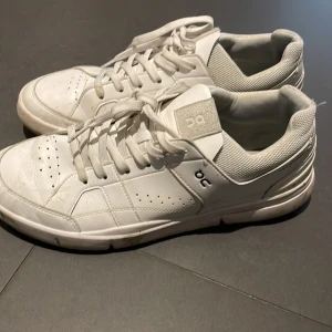 Vita sneakers från ON - Säljer ett par vita sneakers från ON med klassisk snörning och perforerade detaljer för ventilation. Skorna har en stilren design med märkets logotyp på sidan och baksidan. Perfekta för en avslappnad stil.