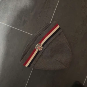 Grå mössa med randigt band - Snygg grå mössa med ett randigt band i rött, vitt och blått. Perfekt för kyliga dagar. Märkeslogga framtill ger en extra touch. Passa på o skriva vid minsta funderingen,priset går att diskutera 