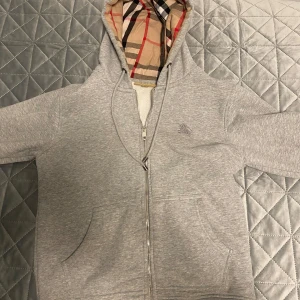 Grå hoodie från Burberry - Säljer en stilren grå hoodie från Burberry med dragkedja och klassiskt rutigt foder. Hoodien har en broderad logga på bröstet och är perfekt för en avslappnad look. Den har långa ärmar och en mysig känsla. Perfekt för kyliga dagar! Pris går självklart att diskutera, var öppen med frågor och prisförslag, pris går självklart att diskutera.