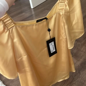 Gul offshoulder topp från Stockh LM - Säljer en snygg gul offshoulder topp från Stockh LM. Toppen har korta ärmar och är tillverkad i ett glansigt material som ger en lyxig känsla. Perfekt för att liva upp garderoben med lite färg!  Kunden står för frakten/ alternativt mötas upp i Norrköping!