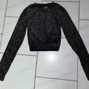 Svart glittrig topp - Snygg svart glittrig topp med långa ärmar och en twist-detalj framtill. Perfekt för en kväll ute eller festliga tillfällen. Toppen har en croppad passform som ger en trendig look.