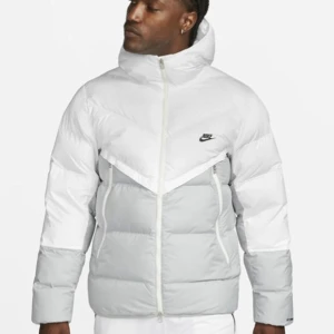 Vit och grå dunjacka från Nike - Säljer en snygg vit och grå dunjacka från Nike. Jackan har en hög krage och dragkedja framtill. Den är perfekt för kyliga dagar och har en stilren design med Nike-loggan på bröstet.