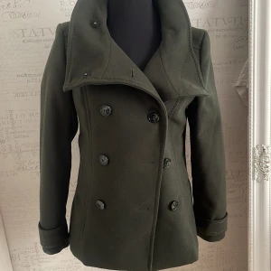 kappa från H&M - Snygg olivgrön kappa från H&M med dubbelknäppning och bred krage. Jackan har långa ärmar och en stilren design med knappar både fram och bak. Perfekt för kyligare dagar. Nyskick