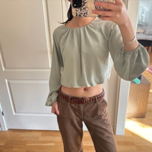 Grön croppad flowy blus - Säljer en stilren grön croppad blus med långa ärmar och elastisk halsringning. Perfekt för en avslappnad look.