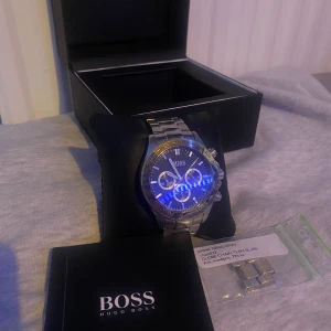 Silverfärgad klocka från Hugo Boss - Snygg och stilren klocka från Hugo Boss med silverfärgad länk och svart urtavla. Klockan har tre sub-dials och en tachymeter på ringen. Perfekt för den som vill ha en elegant accessoar. Vatten tät och inte några skråmor. 