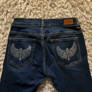 Vintage flare lowrise jeans - Snygga mörkblå jeans med låg midja och unika vingdetaljer i silver på bakfickorna. Perfekta för dig som vill ha något extra i garderoben. De har en klassisk femficksdesign och stängs med knapp och dragkedja. Midjemåttet är ca 35cm och Innerbenslängden ca 78 cm. Priset går att pruta!