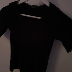 Svart topp med spetsdetaljer från monki  - Snygg svart topp med spetsdetaljer vid halsringning och ärmslut. Perfekt för en stilren look. Toppen har en elegant design som passar till många tillfällen.