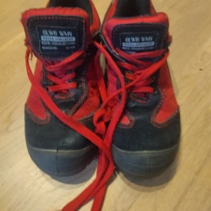 Röda och svarta skor med snörning - Säljer ett par robusta skor i rött och svart med snörning. Skorna har en slitstark sula och är perfekta för utomhusaktiviteter. Märket är Oliva Wan, vilket ger en unik stil.