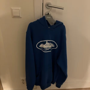 Blå Corteiz hoodie - Säljer en blå hoodie med ett vitt tryck på framsidan. Tröjan har en klassisk design med huva och långa ärmar. Perfekt för en avslappnad stil.