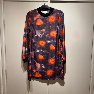 Mönstrad klänning från Monki strl M - Snygg klänning från Monki med ett färgstarkt mönster i orange och lila toner. Klänningen har långa ärmar och en rund halsringning med svart ribbad kant. Perfekt för att sticka ut med stil!
