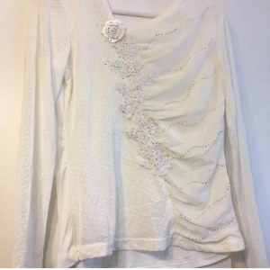 Vit långärmad blus med spetsdetaljer storlek S/M - Elegant vit blus med långa ärmar och vackra spetsdetaljer. Blusen har en unik design med draperade veck och en dekorativ blomma på framsidan. Perfekt för en stilren look.