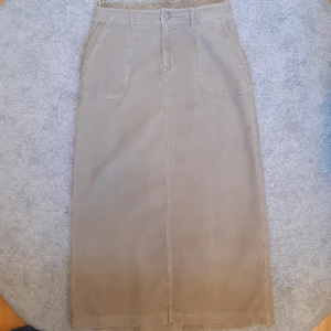 Lång manchesterkjol - Lång beige manchesterkjol med slits baktill. Kjolen har fickor fram och bak samt en knapp och dragkedja i midjan. Perfekt skick!