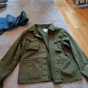 Grön jacka från Gina Tricot - Säljer en grön jacka från Gina Tricot med flera praktiska fickor och knappar framtill. Jackan har en klassisk krage och långa ärmar, perfekt för en avslappnad stil. Passar bra till både jeans och kjol. 🌿 Den har även lite fläckar just här på bilderna men den ska såklart tvättas! 