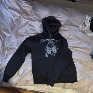Svart hoodie från Abundant brands - Säljer en svart hoodie med dragkedja och rhinestone som bildar texten 'ABUNDANT' och ett dekorativt mönster. Perfekt för en unik och stilren look. Hör av dig vid intresse!