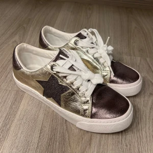 Glittriga sneakers från Next - Snygga sneakers i glittrigt guld och brunt från Next i storlek 38,5. Slutsålda på zalando. Skorna har en stjärndetalj på sidan och vita snören. Skorna har endast andvänts en gång!!