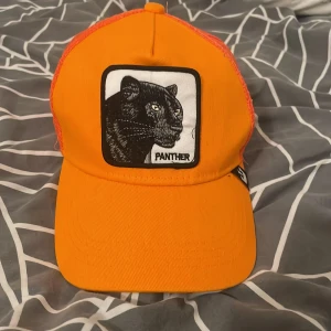 Orange keps goorin bros med panther tryck - Snygg orange keps med ett coolt pantermotiv på framsidan. Kepsen har ett nätmaterial på baksidan för extra ventilation och en justerbar rem för perfekt passform. Perfekt för en avslappnad stil.