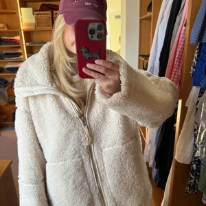 Vit teddyjacka med dragkedja - Mysig vit teddyjacka med dragkedja framtill. Jackan har en hög krage och är perfekt för kyligare dagar. Den fluffiga texturen ger en trendig look och den är lätt att matcha med olika outfits.