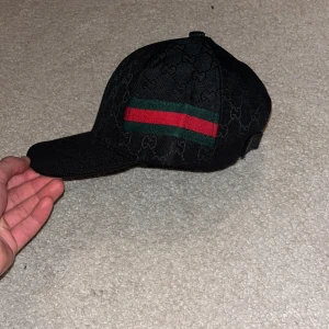 Svart keps med ränder - Snygg svart Gucci keps med diskret mönster och breda ränder i rött och grönt. Perfekt för en stilren look. Funkar med allt och skicket är 10/10 då den aldrig använts